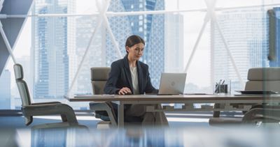 A woman in a suit is sitting in front of a laptop in a large glass office