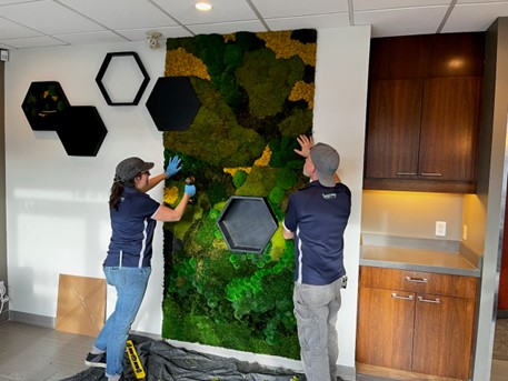 Two people in blue t-shirts and grey baseball caps are installing a plant walll
